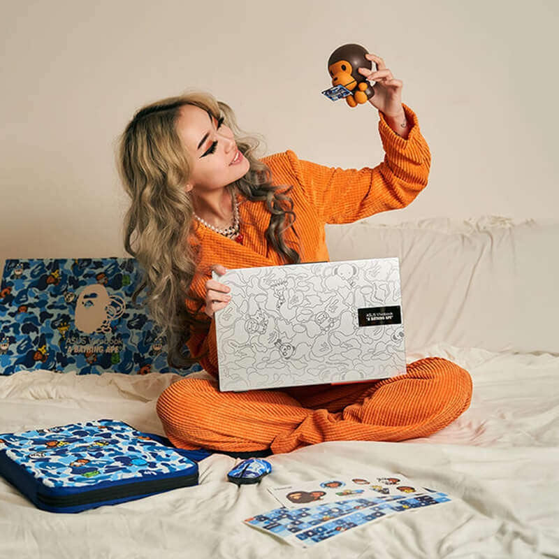 A woman dressed in an orange suit is sat cross legged on a bed with ASUS Vivobook BAPE® Edition bundle elements spread out beside her. She is holding a silver laptop in her right hand and a figurine in her left.