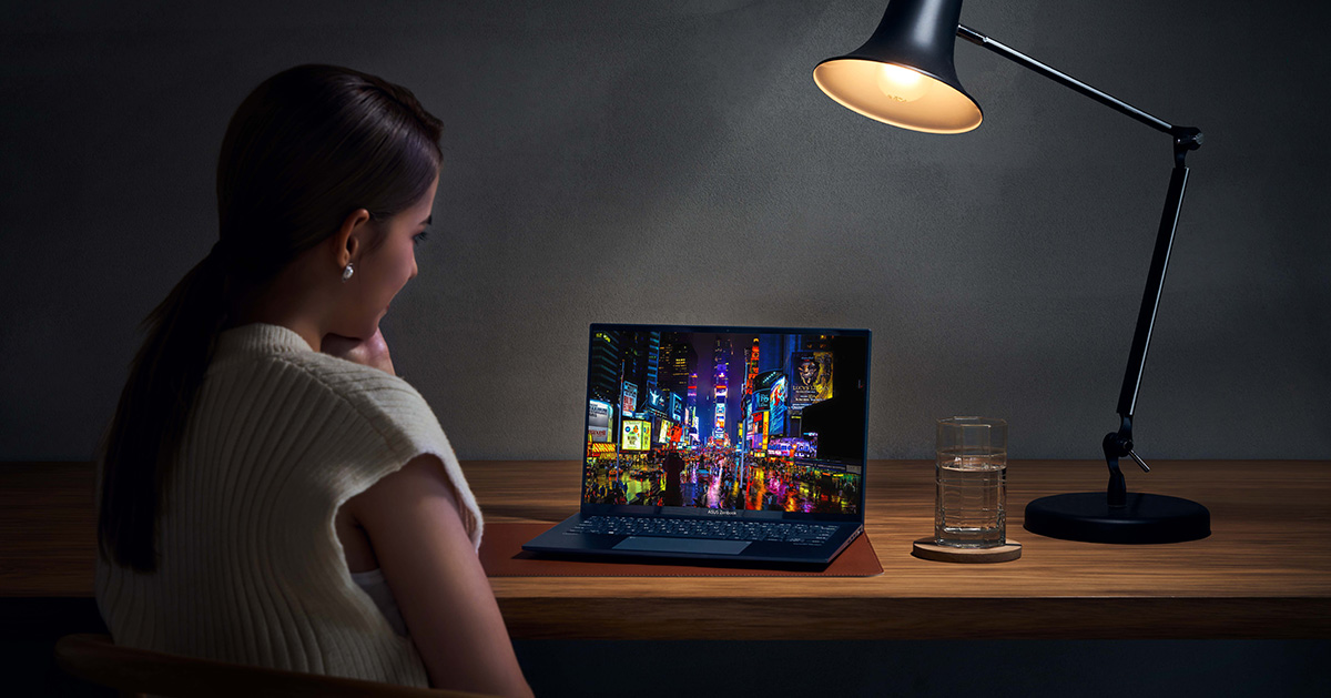 A woman watches a movie by a laptop in dark environment. The only light comes from the lamp on the table. The OLED screen displays a vivid city night view.
