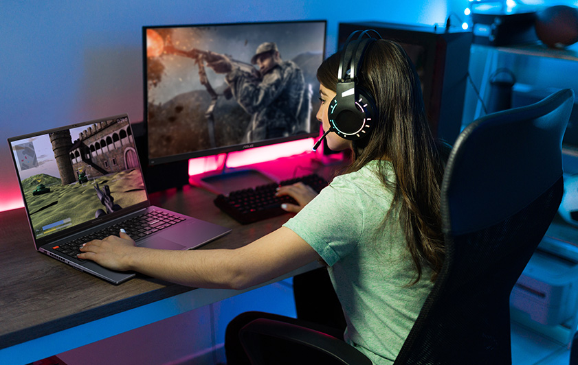 A girl with headphone and microphone is playing online game by both laptop and desktop simultaneously.