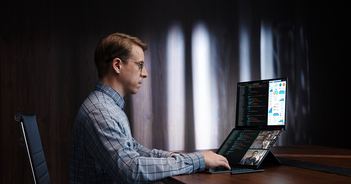 mężczyzna siedzący przy biurku i korzystający podczas pracy z dwuekranowego laptopa ASUS Zenbook DUO. Laptop wyświetla różne okna na obu ekranach, natomiast na dolnym ekranie prowadzona jest wideokonferencja