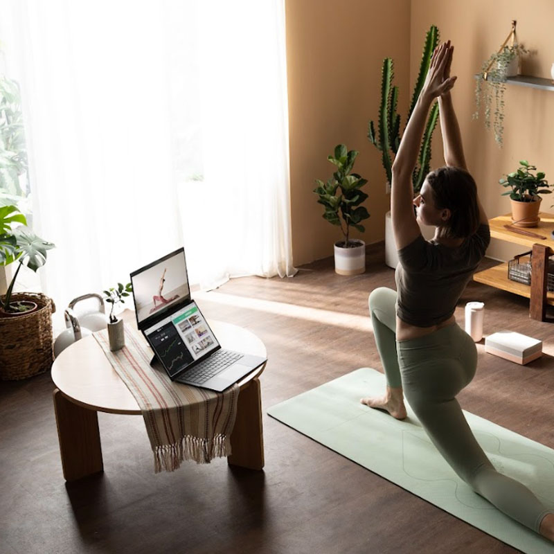 Kobieta na jasnozielonej macie do jogi ćwicząca jogę po prawej stronie. Patrzy na laptopa z dwoma ekranami, postawionego na okrągłym stole po jej lewej stronie. Laptop jest wyposażony w dwa ekrany. Ekran górny wyświetla samouczek jogi tej kobiety, natomiast ekran na dolny jest podzielony na dwie części i wyświetla różne informacje.