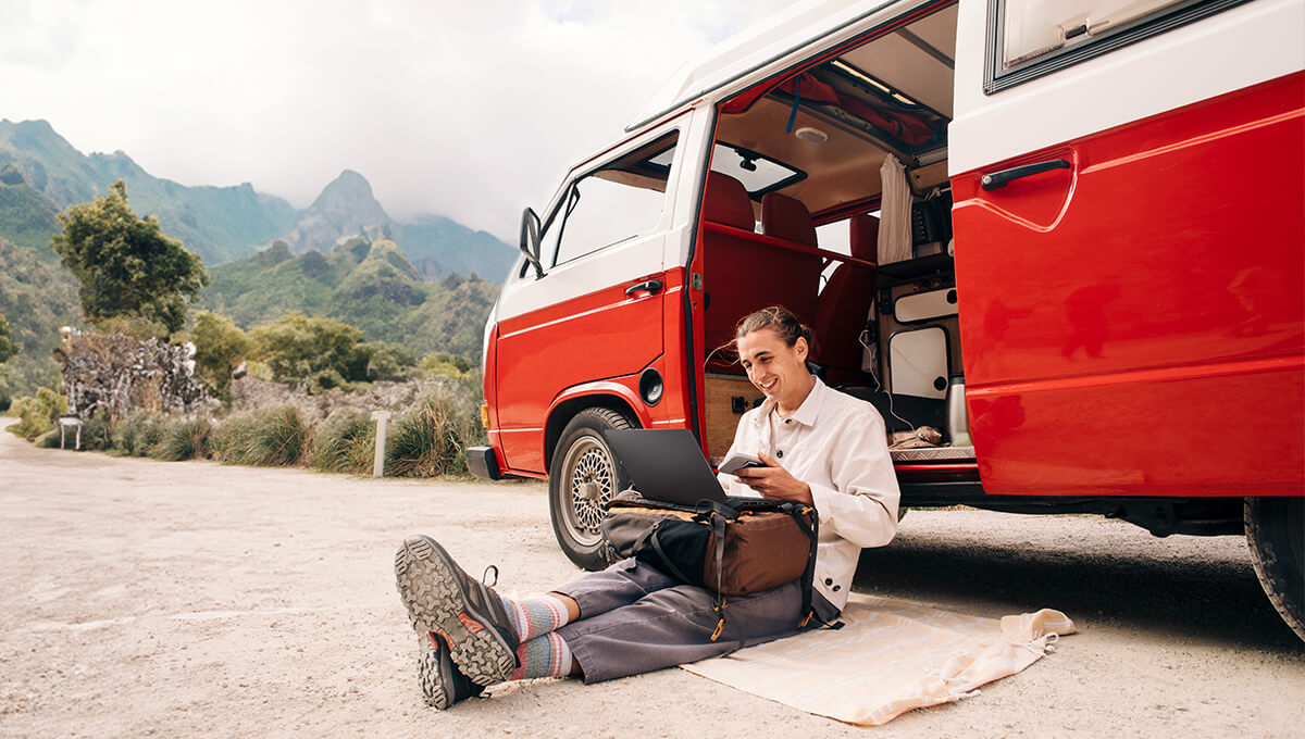 A man is sitting on a dirt road where is red and white van is parked. He has his ProArt laptop propped up on his backpack and is smiling while using it.
