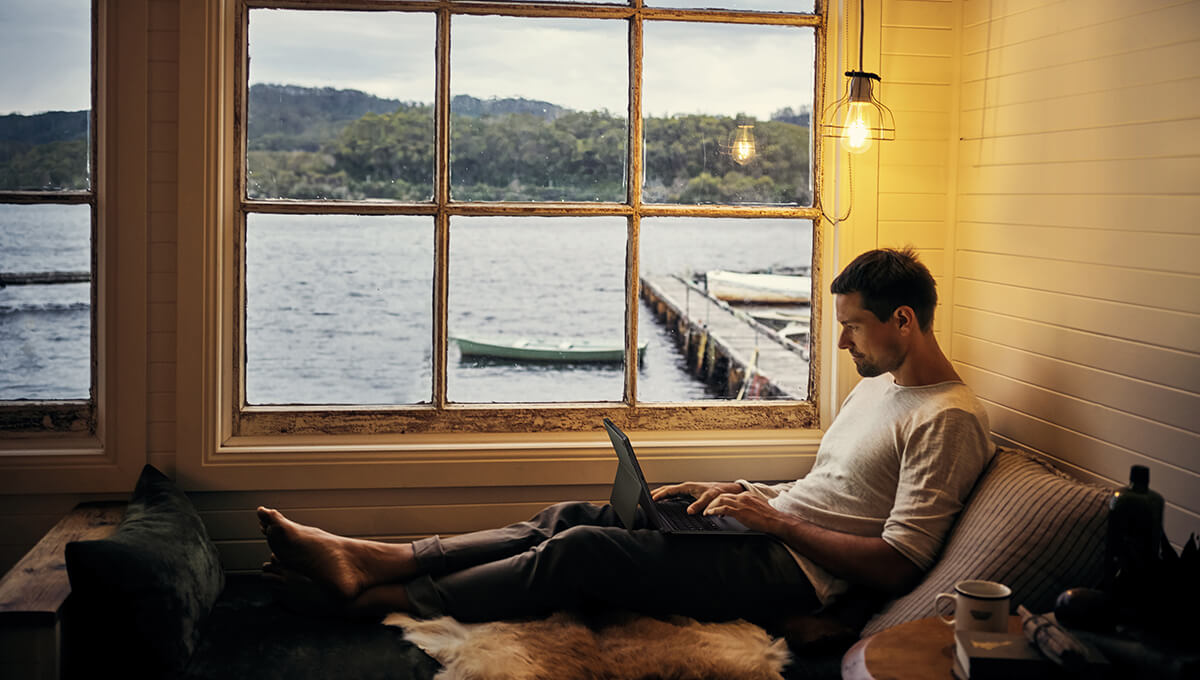 A man is sitting by the window with his legs stretched out in front of him and a detachable laptop placed on his lap. Outside the window there is a dock with a small fishing boat tied to it.
