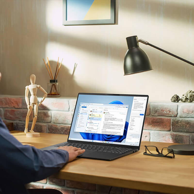 Een man in een blauw shirt gebruikt Copilot-functies op een ASUS-laptop op een houten bureau.