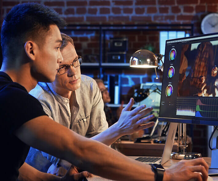 Twee mannen zijn geconcentreerd bezig met het color grading van een video op een ProArt-monitor, in een warm verlicht kantoor met bakstenen muren, en stralen samenwerking en concentratie uit.