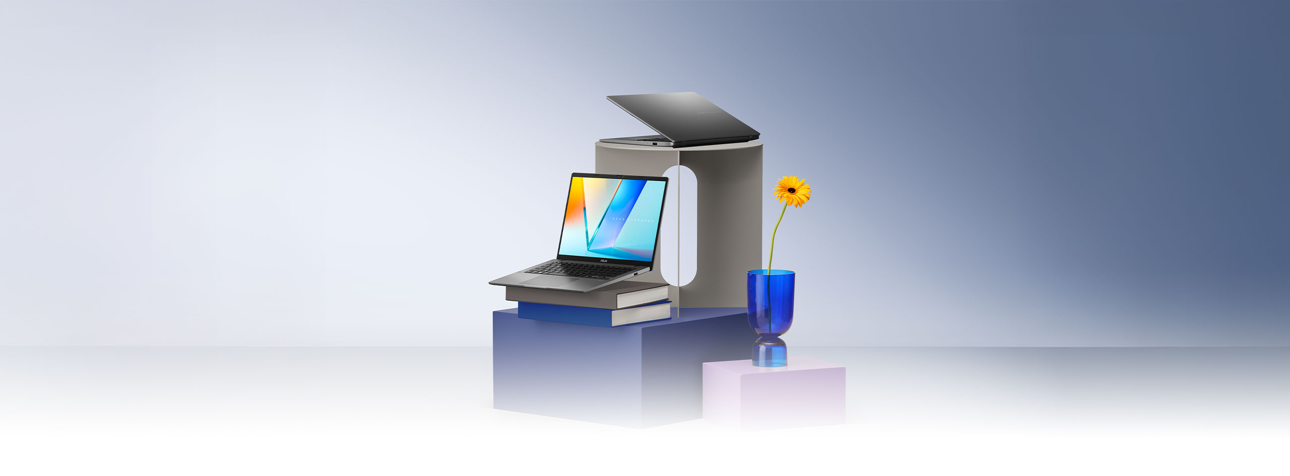 Two open laptops, one gray and one silver, are displayed on stacked gray, blue, and purple pedestals, with one resting on a pair of books. A single yellow flower in a blue glass vase stands nearby.