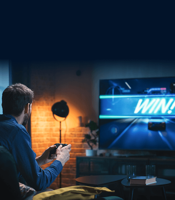 A man sits on a couch, holding a game controller and facing a large screen displaying a racing game with the word 'WIN!'.