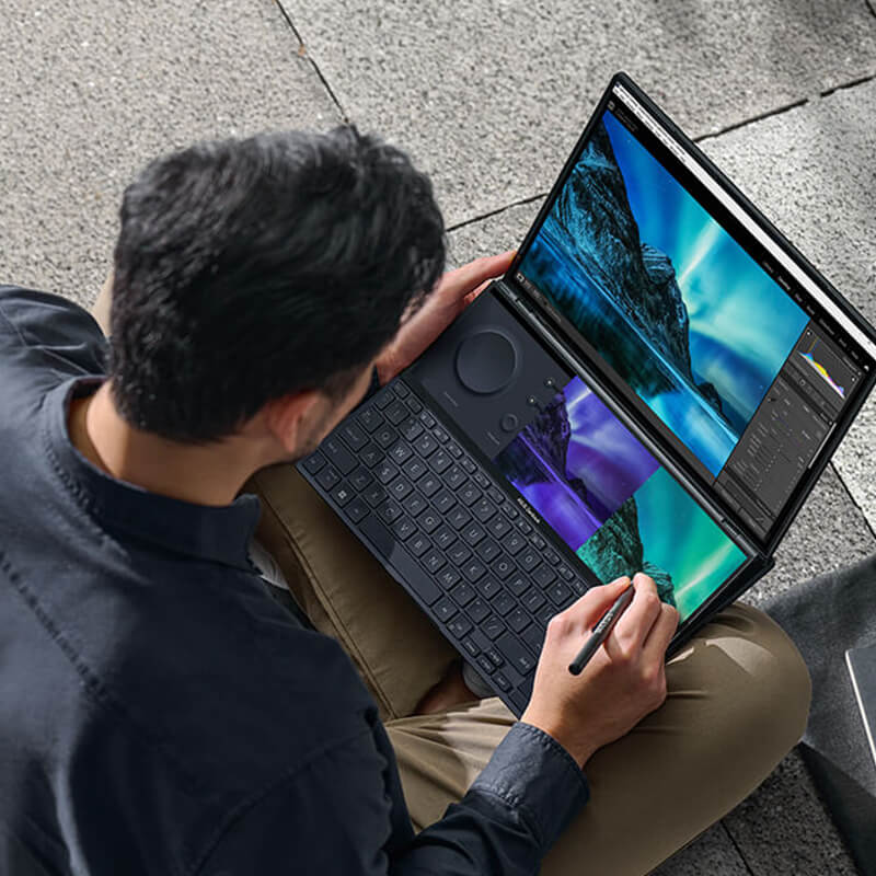 a top view of a young man using a stylus on the ASUS Screenpad plus of the Zenbook Pro Duo 14 OLED dual screen laptop while sitting on stairs outdoors
