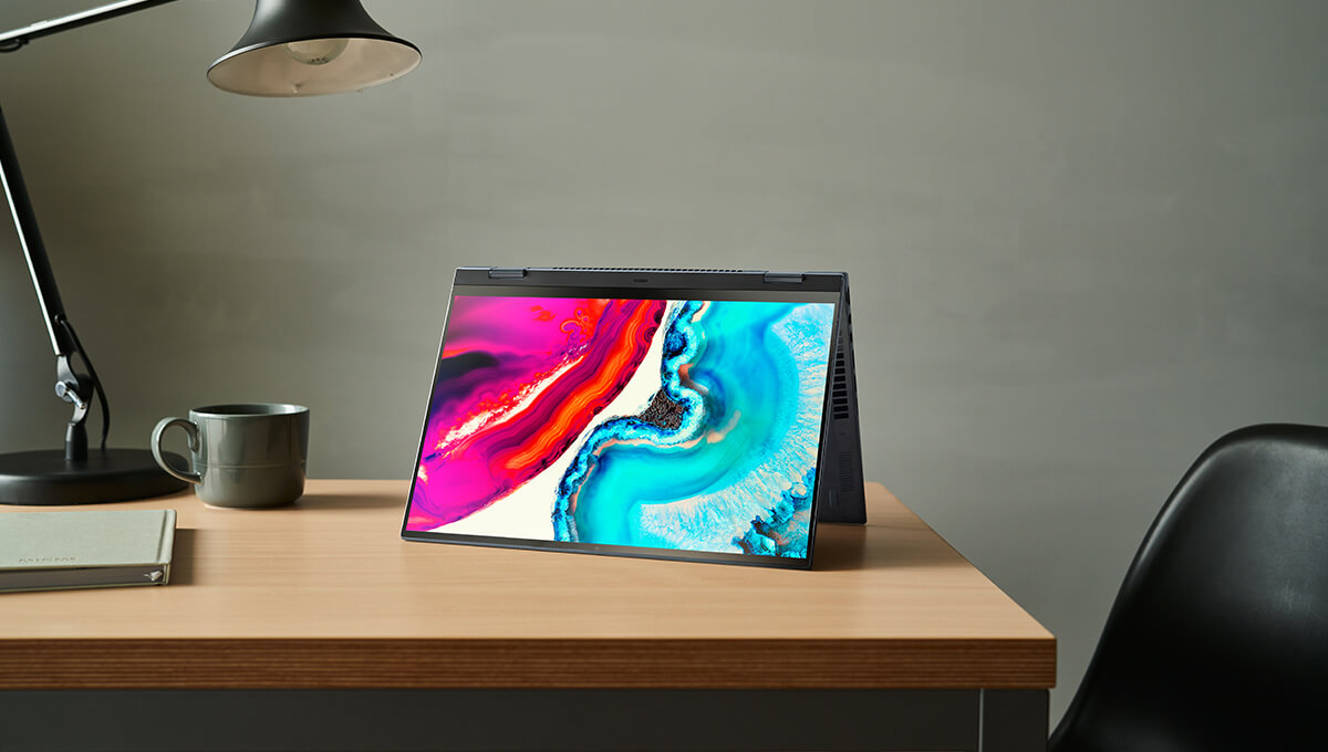 A Zenbook laptop is placed on the table next to a lamp closed and a cup. The laptop screen, which is ASUS Lumina OLED display shows complementary colors, with rose red on the left side and aqua on the right.