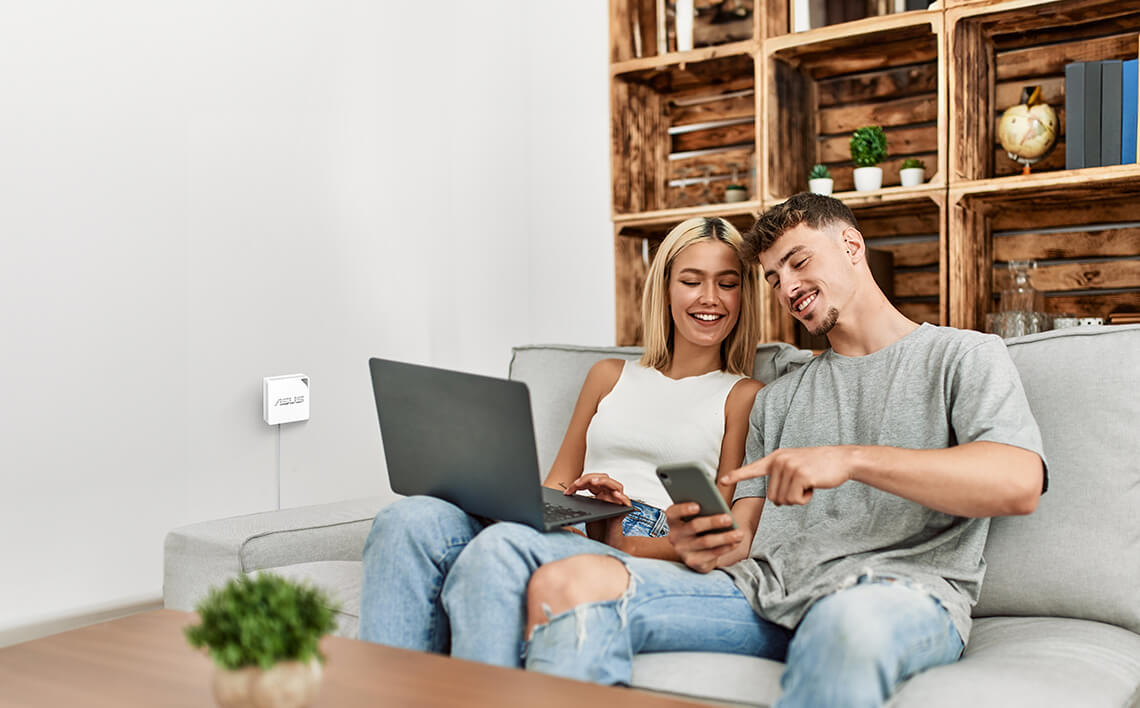 A smiling young couple sits on the sofa at home, using laptop and smartphone together.