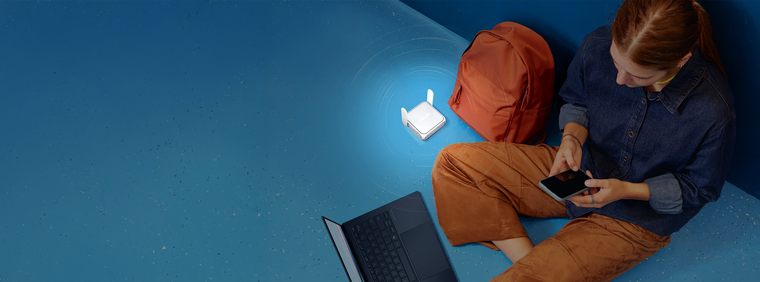 Une femme est assise sur le sol et utilise un smartphone et un ordinateur portable. L'ASUS RT-BE58 Go est placé à proximité, à côté d'un sac à dos.