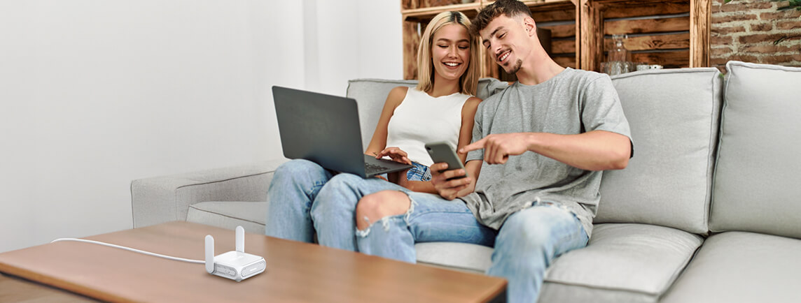 Un jeune couple assis sur un canapé à la maison, utilisant ensemble un ordinateur portable et un smartphone, avec l'ASUS RT-BE58 Go sur la table devant eux.