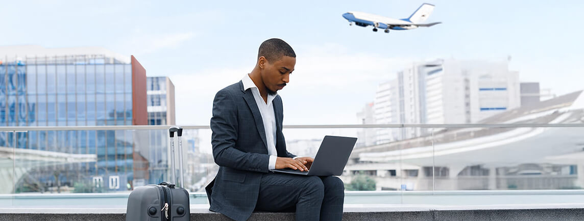 Un homme d'affaires travaillant sur un ordinateur portable à l'aéroport, avec une valise à côté de lui et un avion volant au-dessus de sa tête.