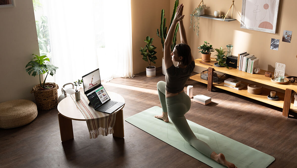 A la derecha, una mujer hace yoga sobre un tapete verde claro. Ella está mirando una laptop de doble pantalla apoyada en la mesa redonda a su izquierda. Hay dos pantallas en la laptop. La de arriba muestra el tutorial de yoga de la mujer, mientras que la de abajo está en pantalla dividida y muestra información diferente.