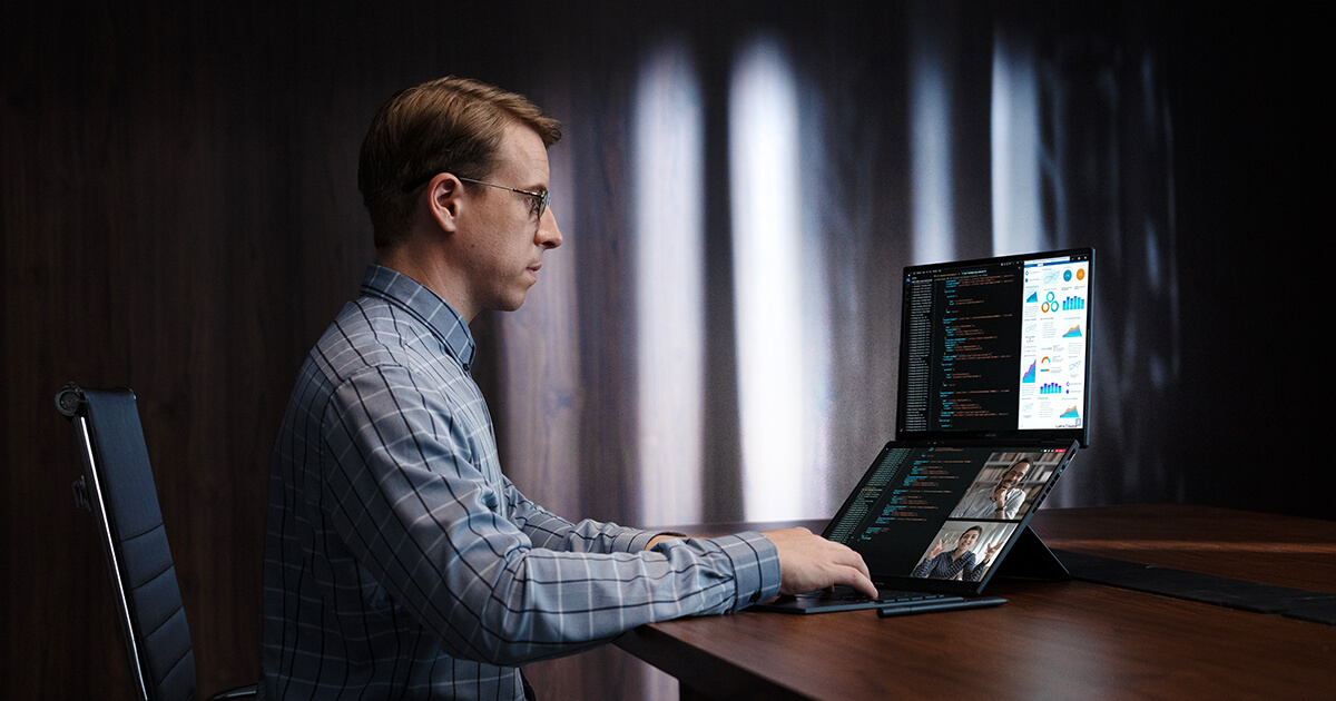 Un hombre vestido con ropa de trabajo utiliza una laptop de doble pantalla sobre una gran mesa de madera. Está codificando en una mitad de la laptop y consultando e investigando en la otra mitad.