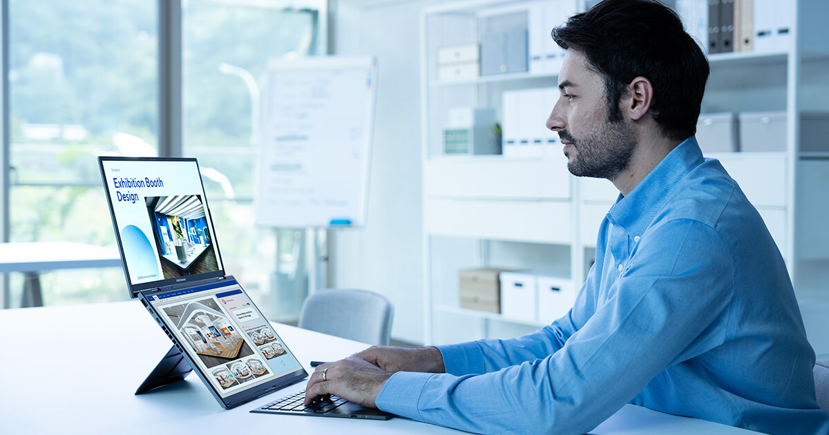 Un hombre con camisa azul está escribiendo en una laptop de doble pantalla a través de un teclado inalámbrico. Está mirando gráficos del diseño de una cabina en la pantalla superior mientras ve referencias en la pantalla inferior