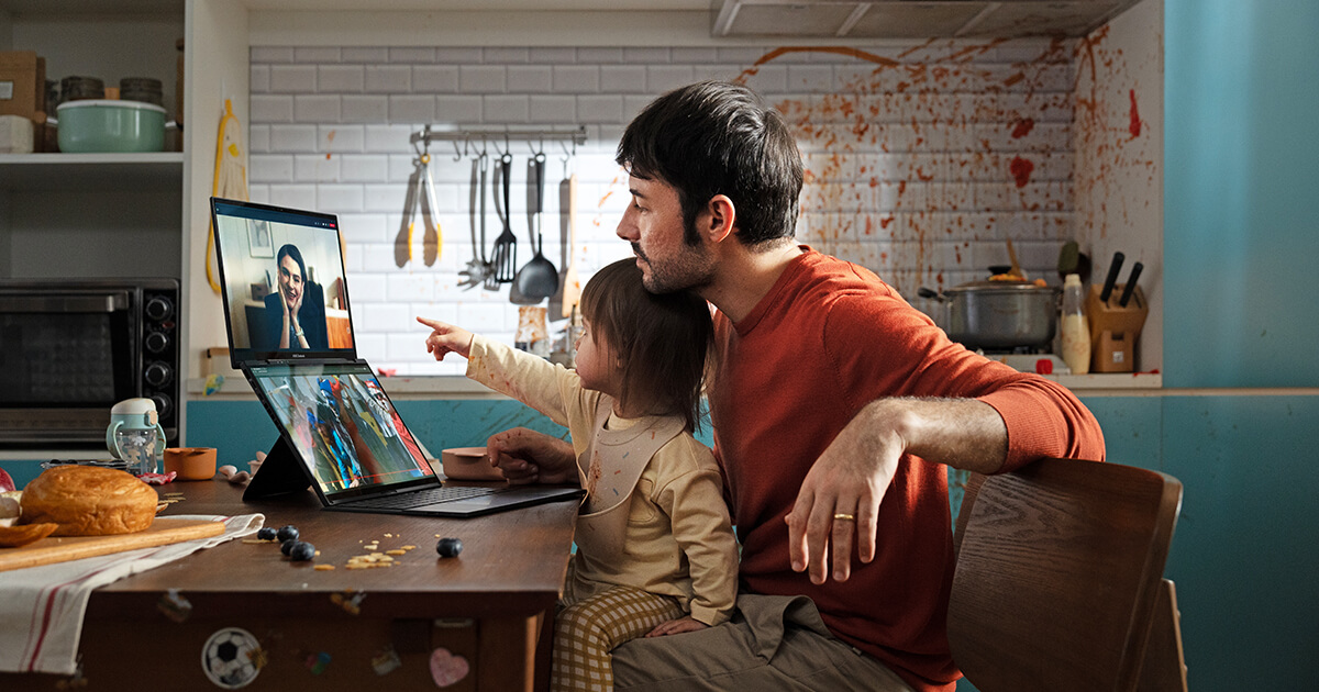 Un padre tiene a su hija sentada en su regazo junto a la mesa de la cocina. Están hablando con una mujer en la pantalla superior de la laptop de doble pantalla colocado frente a ellos y viendo un vídeo en la pantalla inferior.