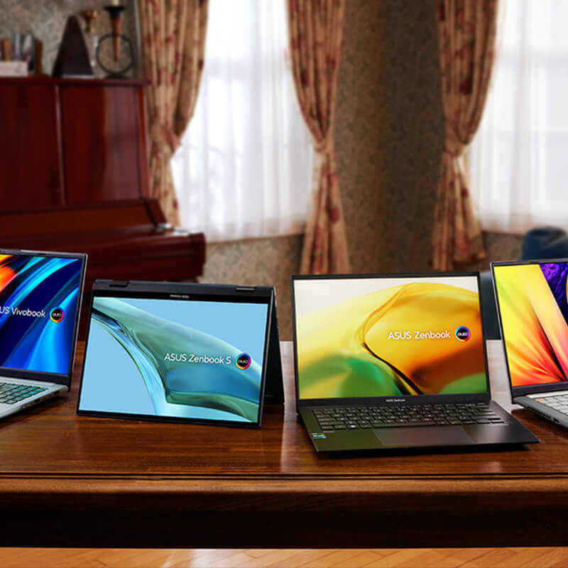 Four ASUS laptops are placed on a wooden table side by side in a living room lit with warm yellow light.