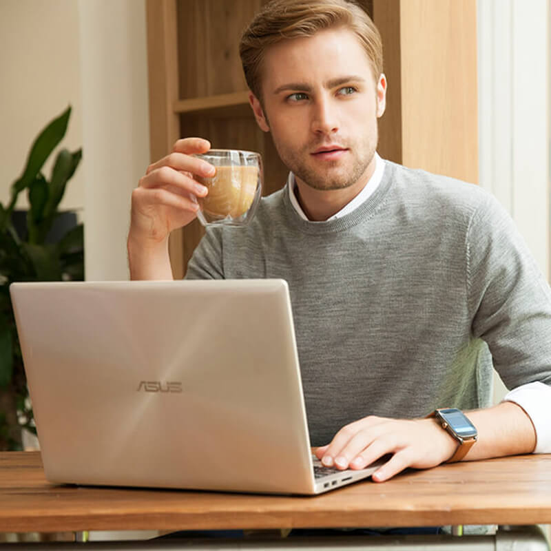 A man puts his right hand on ASUS laptop on the table. He holds a cup of coffee by the other hand and takes a break for a while with his eyes watch outside.