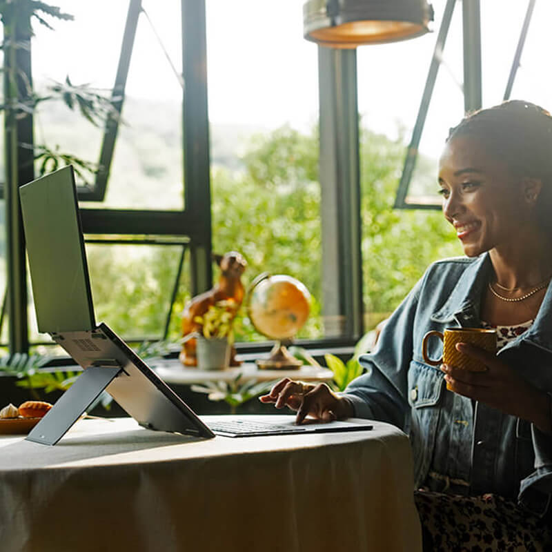 A young woman using ASUS Zenbook DUO in Dual-Screen mode with a smile on her face while holding a cup, sitting in a room full of plants.