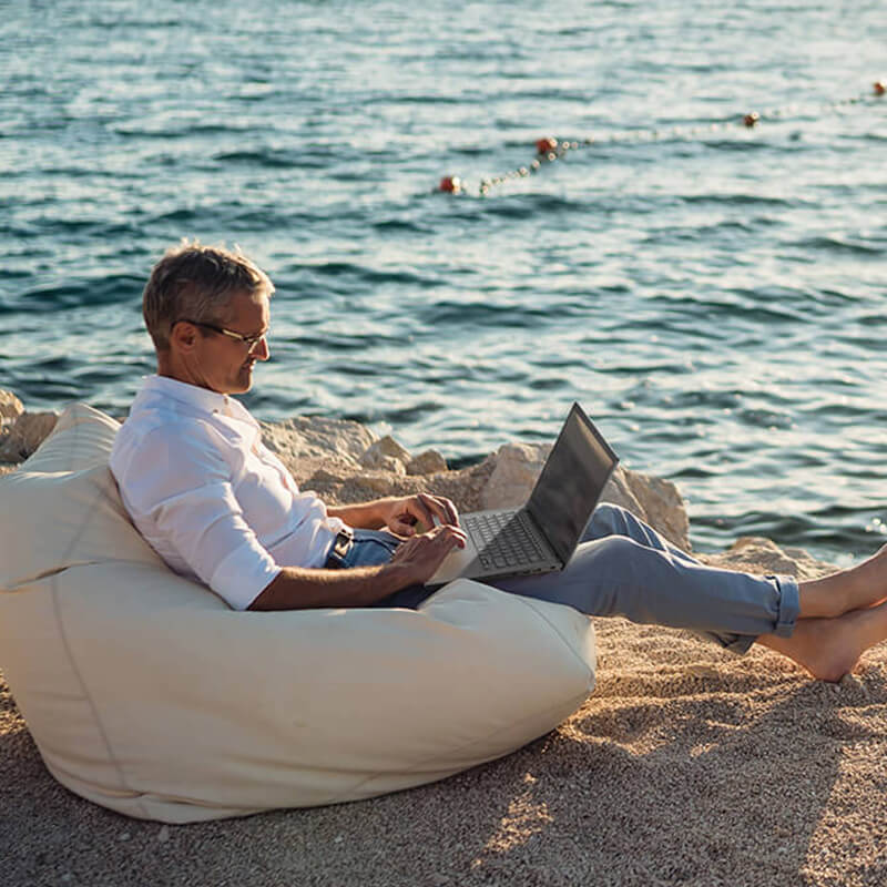 un homme d'âge moyen assis sur une plage, sur un pouf, avec un ordinateur portable ASUS Vivobook sur les genoux
