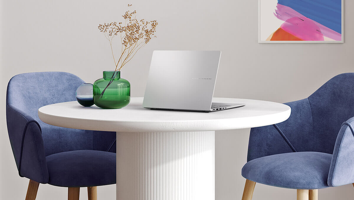 A silver ASUS Vivobook S14 sits on a modern-looking white table with two blue chairs on either side of it.