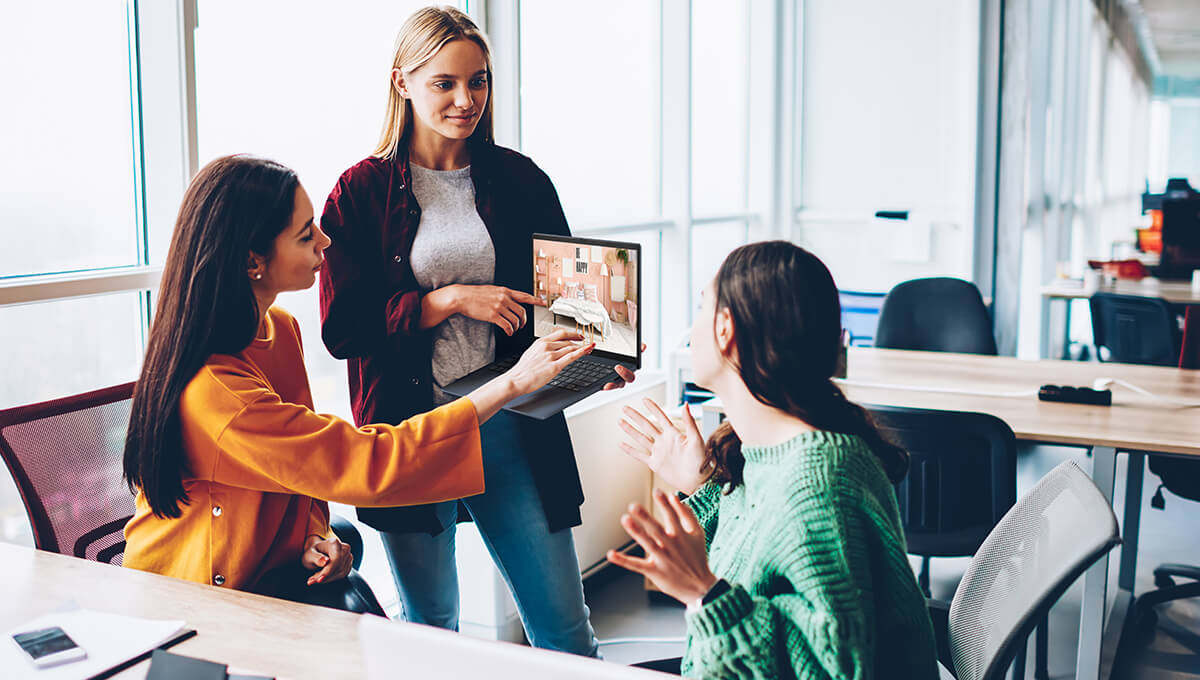 Three girls are discussing in a classroom setting while the girl in the middle holds an ASUS Vivobook 16.