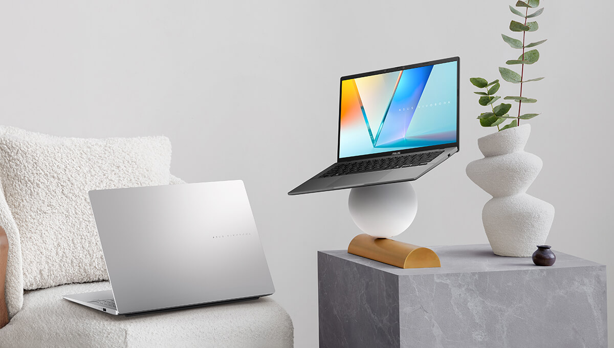 A silver ASUS Vivobook sits on a white couch on the left while a gray ASUS Vivobook is balanced on a white sphere on the right.