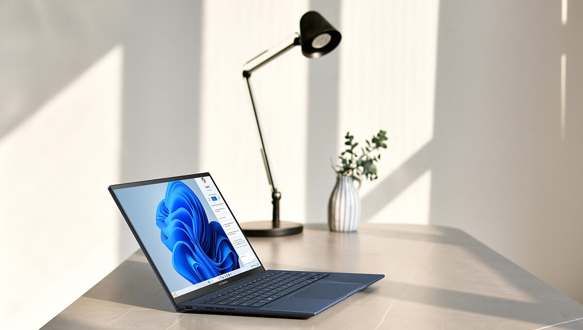 Ноутбук ASUS Zenbook 14 OLED стоит на столе, на его экране виден интерфейс Microsoft Copilot.