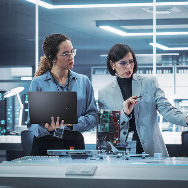Dos mujeres están en un laboratorio discutiendo e investigando. La de la izquierda lleva una camisa azul y sostiene un ordenador portátil ASUS. La de la derecha lleva una americana gris y gesticula a la derecha en medio de la conversación.