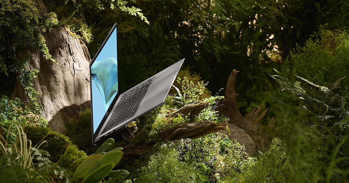 a Zenbook S 13 OLED laptop in Basalt Gray color lying with a screen open inside a sunlit, green forest