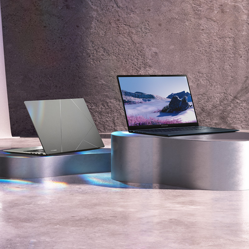 Two Zenbook 15 OLED laptops in Basalt Gray and Ponder Blue colors on metal podiums with dark a concrete wall and a curtain in the background