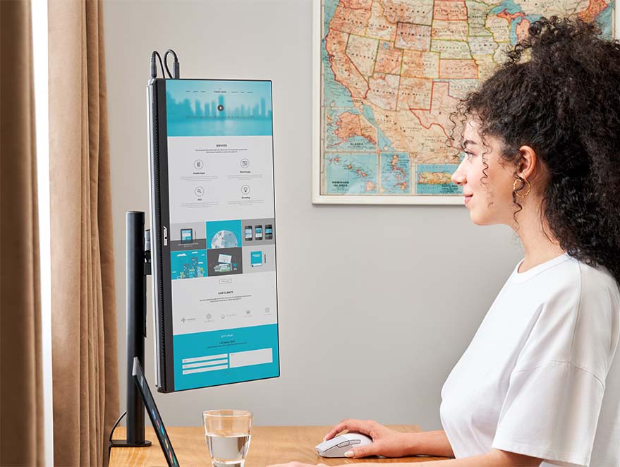 Une femme se tient debout, travaillant à l'aide d'une table de réglage, avec un ordinateur portable et un moniteur portable ZenScreen MB249C, affichant une longue page web en mode portrait.