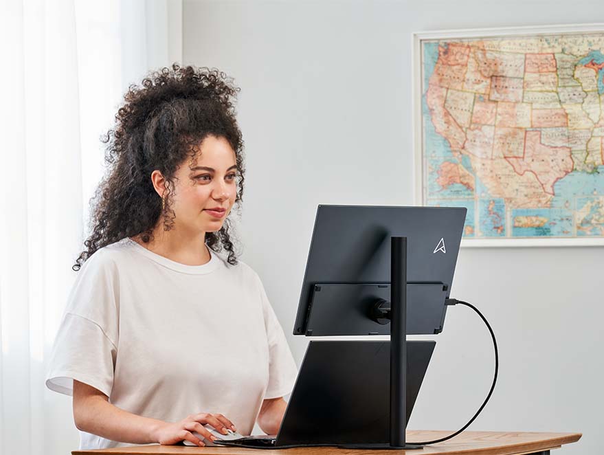 Een vrouw staat te werken aan een verstelbare tafel, met een laptop en de ZenScreen MB16QHG draagbare monitor bevestigd op de MTS02SD standaard, voor een comfortabele werkpositie