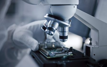 A gloved hand placing an electronic component under a microscope.