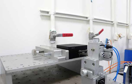 A close-up image of a laboratory setup showcasing a precision mechanical device with a black box, metal clamps, and connected wires against a white background.