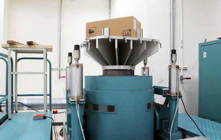 Large industrial vibration testing equipment with a cardboard box on top, surrounded by blue machinery and safety railings.