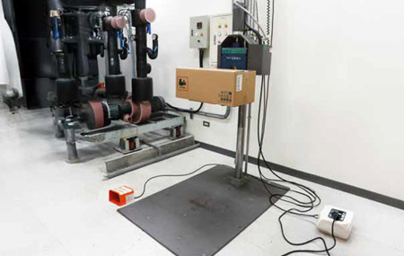 A room with industrial equipment including pipes, control panels, and a cardboard box on a stand, connected with various cables.