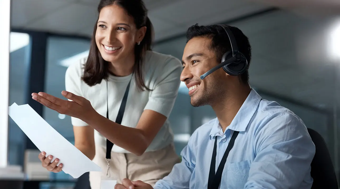 Two office workers discussing, one holding a sheet of paper and the other wearing a headset.