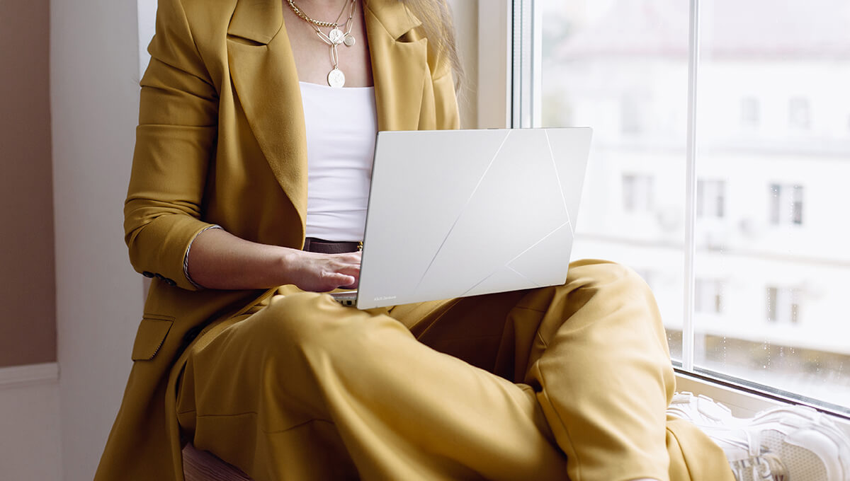 A woman in a yellow suit sits by a window with a Zenbook 14 OLED on her lap.