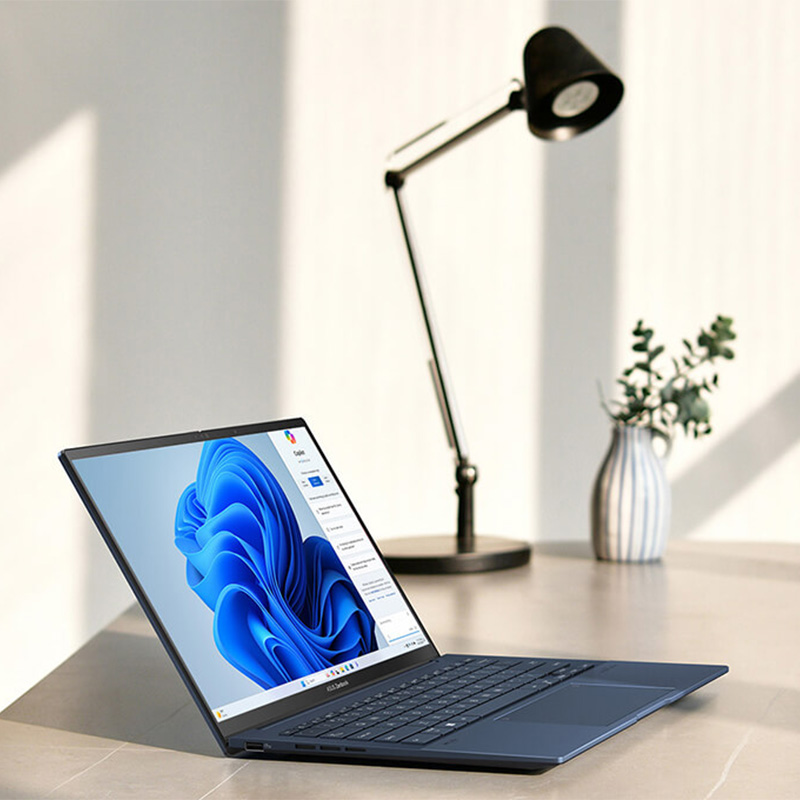 Ноутбук ASUS Zenbook 14 OLED стоит на столе, на его экране виден интерфейс Microsoft Copilot.