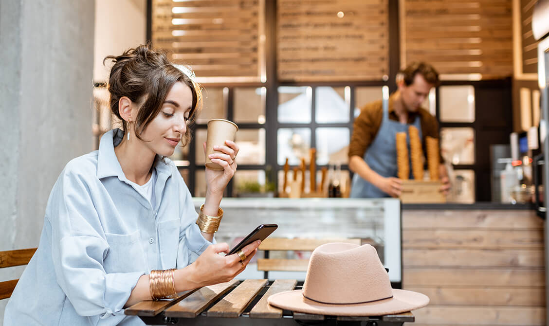 Mladá zákazníčka sedí v kaviarni a používa chytrý telefón, zatiaľ čo v pozadí sa pri pulte venuje práci majiteľ obchodu.
