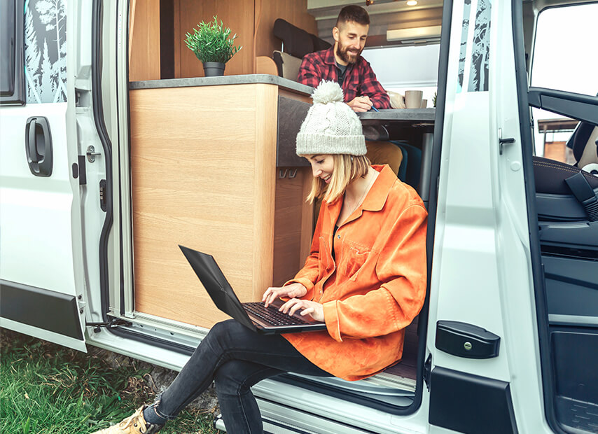 A woman sits at the doorway of a camper van, working on her laptop, while her partner is inside, also engaged in work.