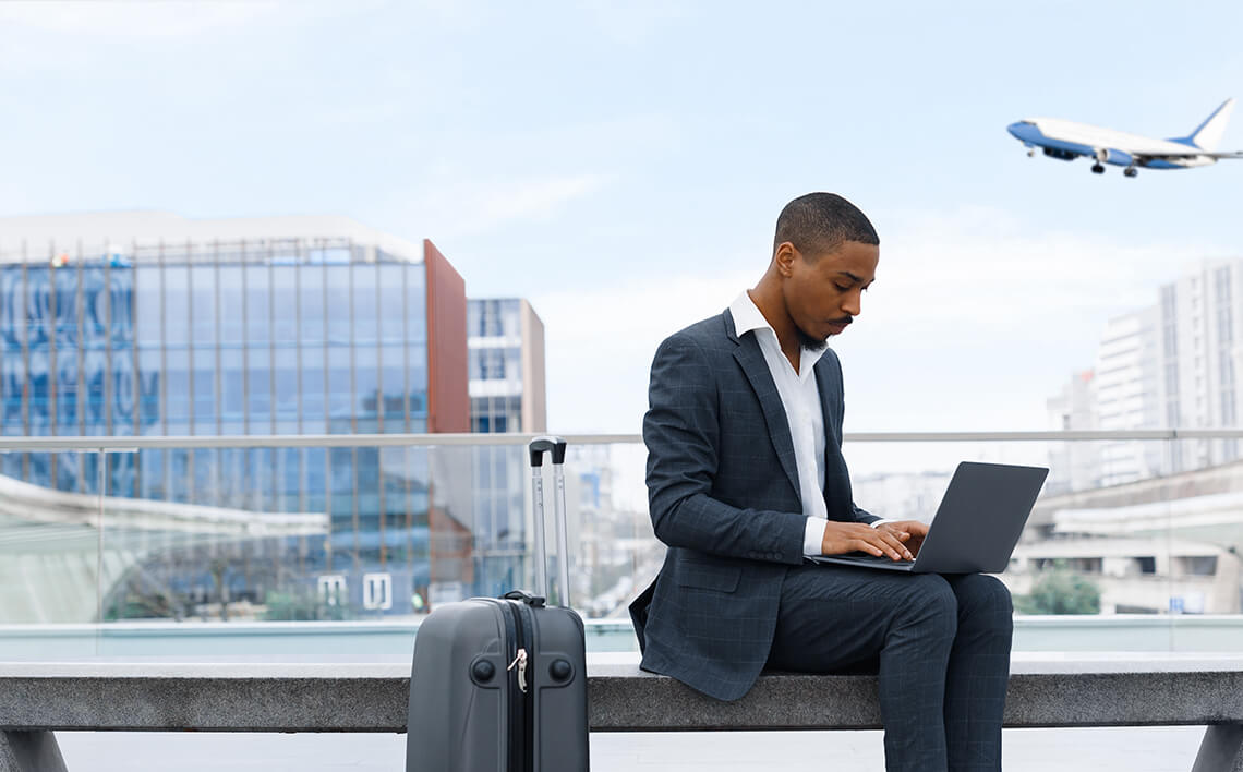 Un hombre de negocios está sentado en un banco del vestíbulo de la terminal, trabajando en su ordenador portátil con una maleta a su lado, mientras un vuelo pasa por el cielo.