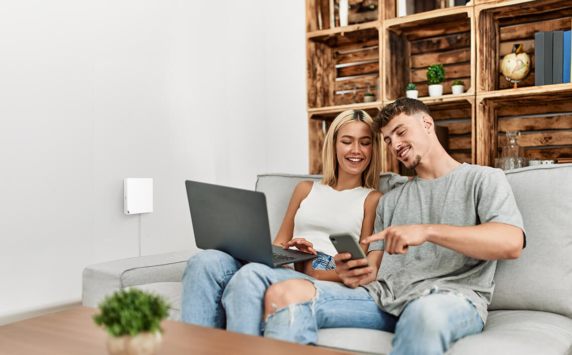 Usmievavý mladý pár sedí doma na pohovke a spoločne používa notebook a telefón.