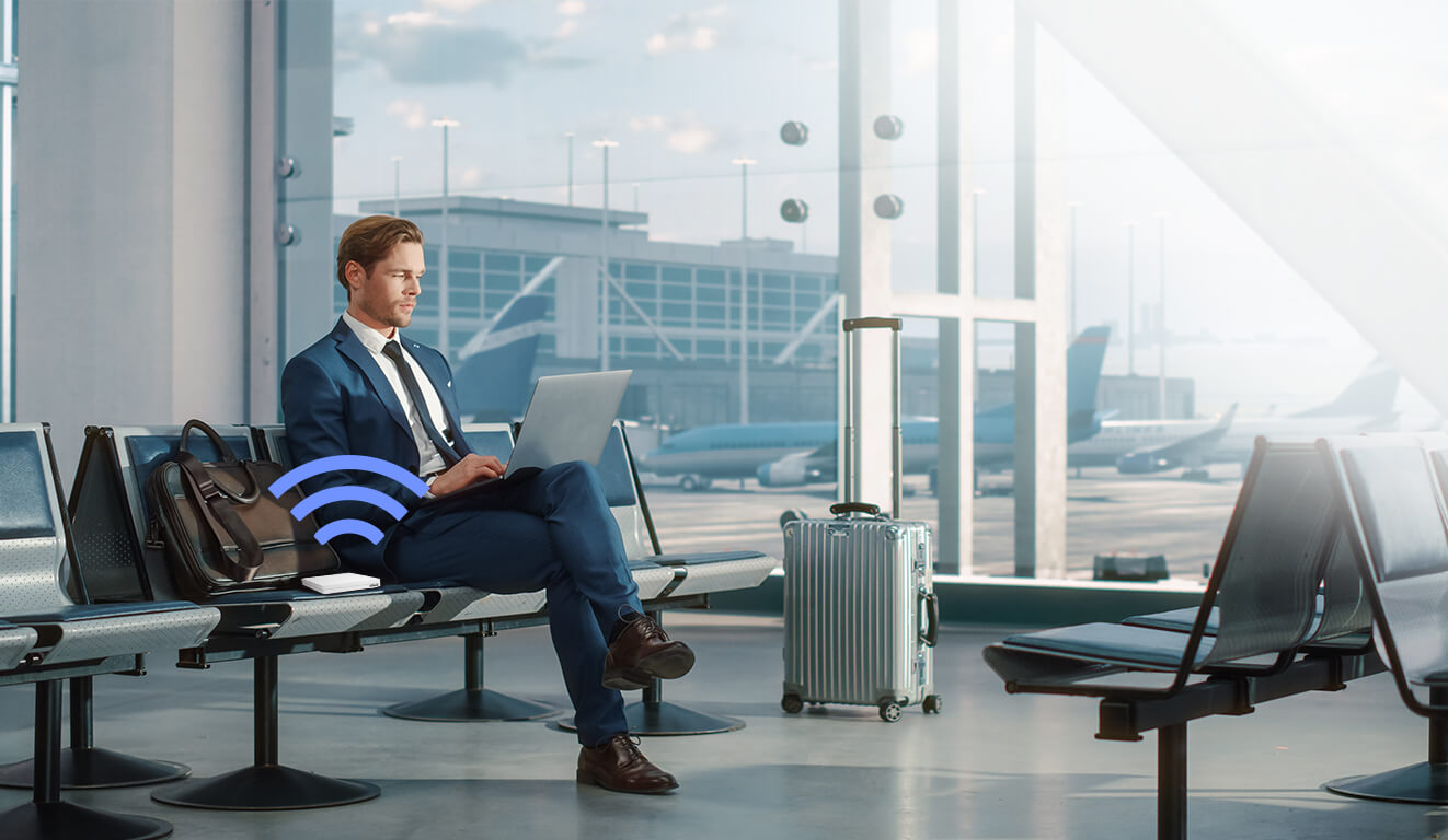 Egy üzletember a reptéren laptopot használva várja a járatát, mellette az RT-AX57 Go router és egy bőrönd.