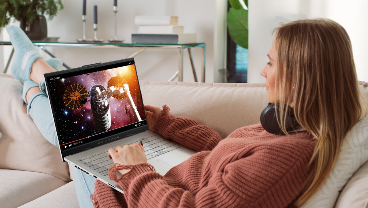 A young woman sitting on a pink sofa with an ASUS laptop on her lap, watching an online video