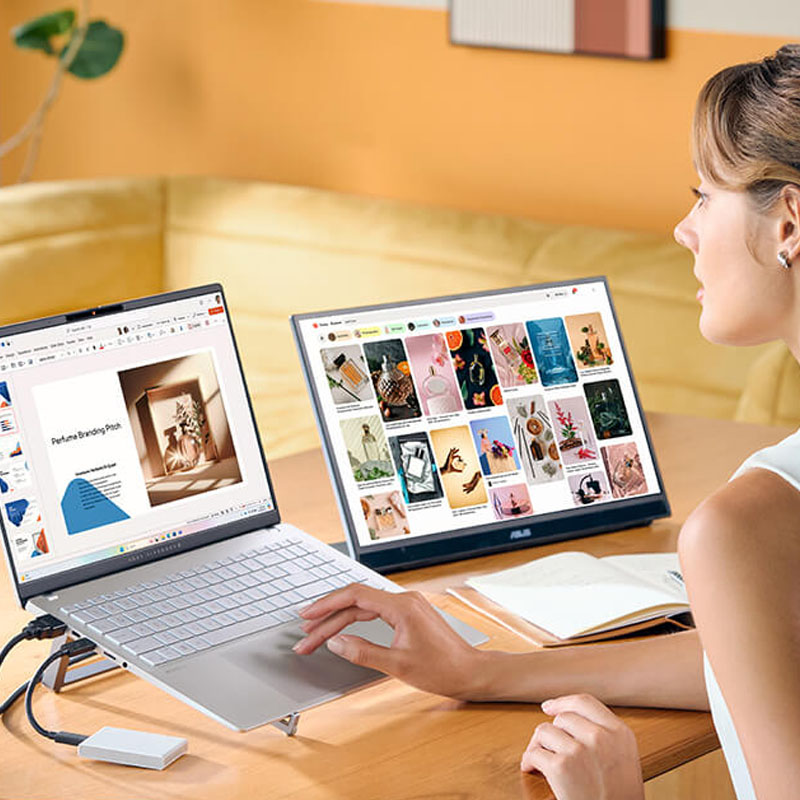 A woman is using a silver ASUS Vivobook on a wooden desk with a screen and a hard drive connected to the laptop.