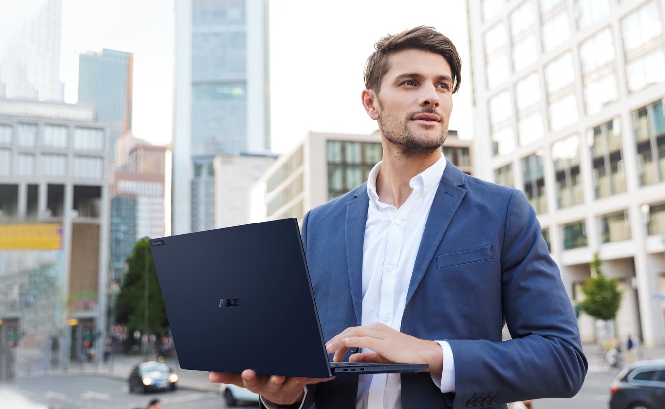 A man in blue business suit working on ExpertBook B5 while walking outdoor, holding the laptop in his right hand.
