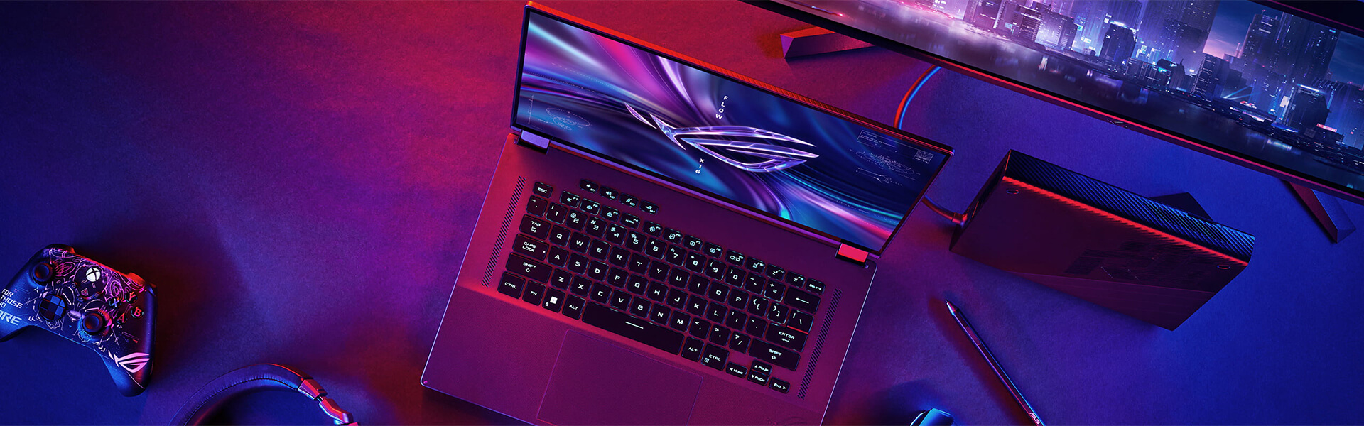 Notebook ROG Flow X16 leží otvorený na stole vedľa herného ovládača, monitora a externej grafiky XG Mobile